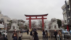 A large tori-i gate protected by a huge stone lion on each side. Through the gate is a walkway lined with cherry trees in bloom.