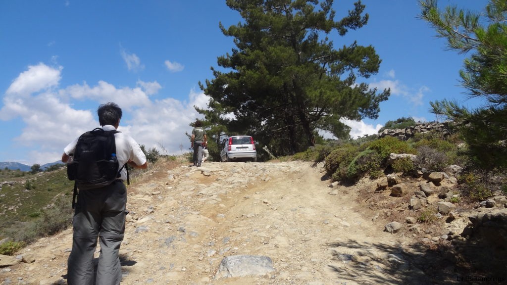 A man wearing a backpack looks up a short incline at the station wagon on top of the dirt path strewn with rocks and ruts.