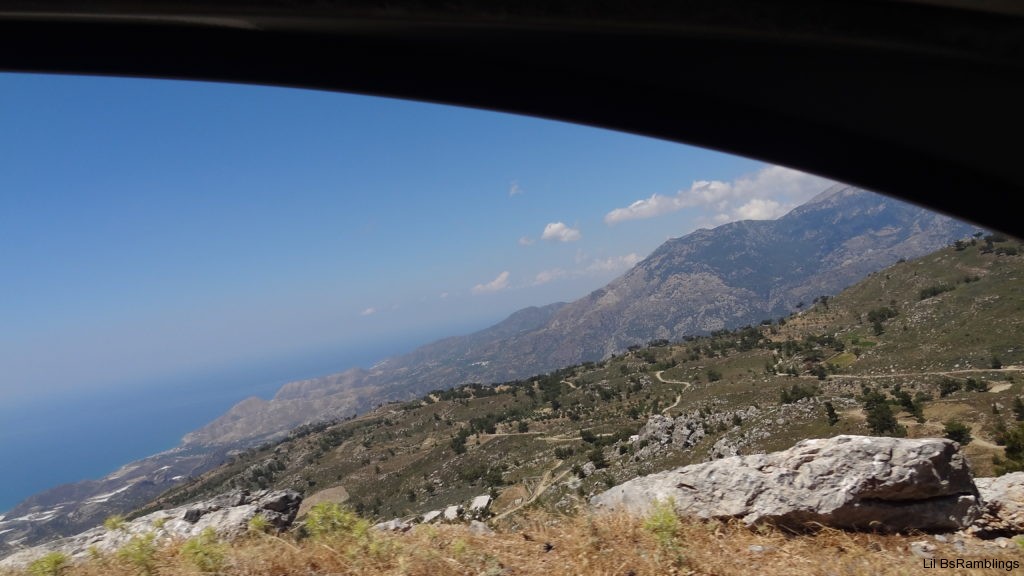 A view of Crete: the dead long grass beside the road leads to a steep rocky drop. Over the rocks you can see a green hill criss-cut with light gray roads and speckled with dark green trees and dark gray boulders. The hazy bluish-brown mountains beyond fade into the ocean to the left and light blue sky dotted with puffy clouds. All that is framed by the upper edge of a car window.
