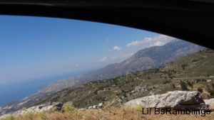 A view of Crete: the dead long grass beside the road leads to a steep rocky drop. Over the rocks you can see a green hill criss-cut with light gray roads and speckled with dark green trees and dark gray boulders. The hazy bluish-brown mountains beyond fade into the ocean to the left and light blue sky dotted with puffy clouds. All that is framed by the upper edge of a car window.