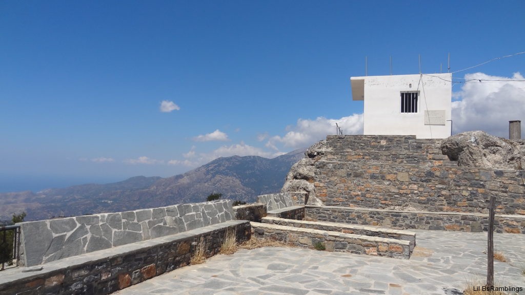 A small white square building sits on a gray stone wall overlooking the mountains of Crete that fade into a faint ocean and vast blue sky as fluffy white clouds roll in from the right.