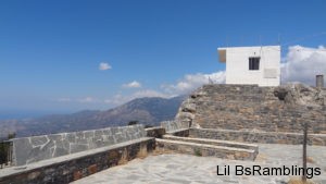 A small white square building sits on a gray stone wall overlooking the mountains of Crete that fade into a faint ocean and vast blue sky as fluffy white clouds roll in from the right.