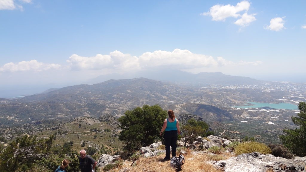 A lady in a blue shirt and dark jeans stands at the edge of a rock outcropping overlooking the majestic mountains of Crete in the background and the city far below to the right.