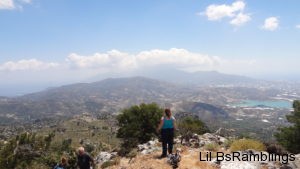 A lady in a blue shirt and dark jeans stands at the edge of a rock outcropping overlooking the majestic mountains of Crete in the background and the city far below to the right.